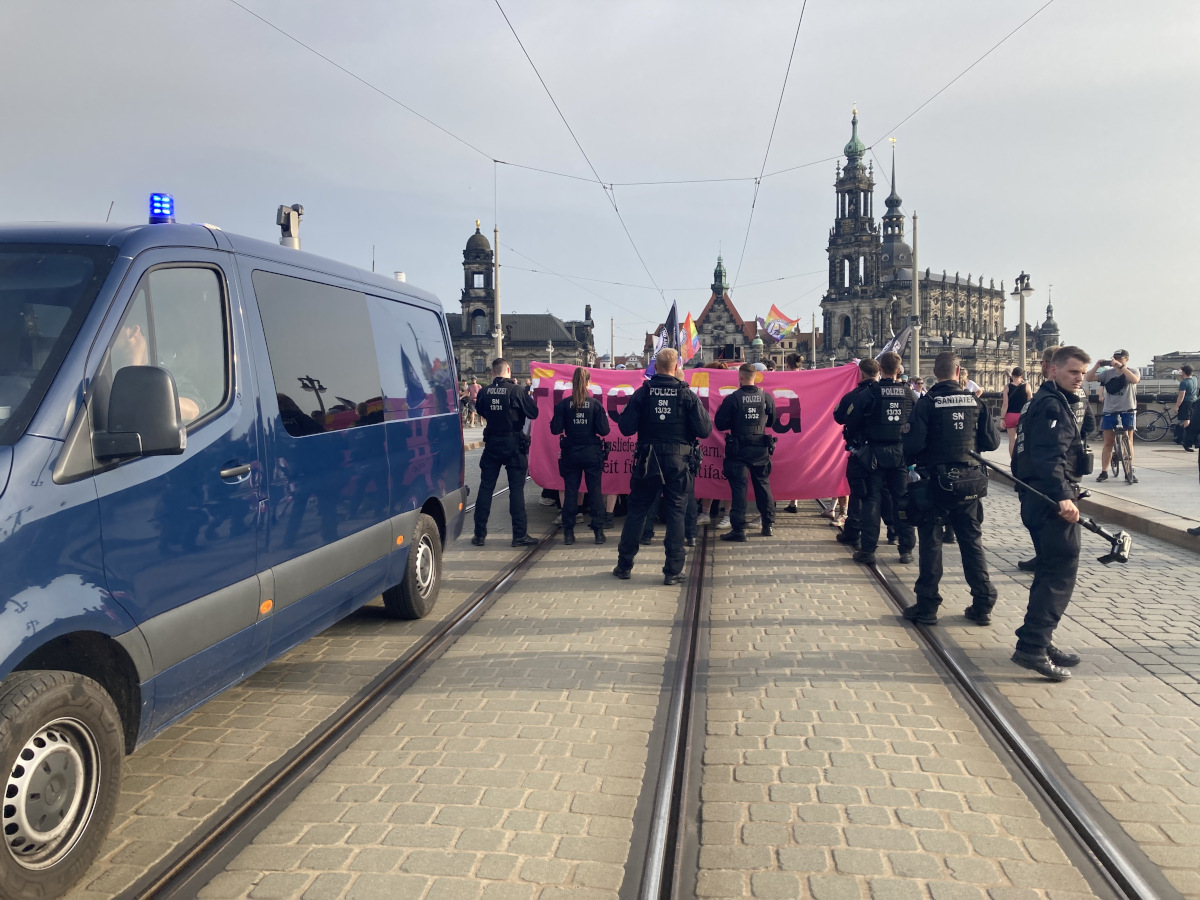 Blick auf die Augustusbrücke in Dresden: Links ein blaues Polizeiauto mit Kamera auf dem Dach, in der Mitte Polizeibeamte vor einer Demonstration mit einem pinken Transparent mit der Aufschrift "Free Maja"